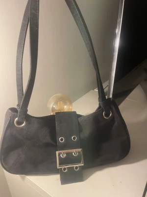 Svart väska med stor bälte-detalj - Snygg svart axelväska med bred bälte-detalj framtill och silverfärgade metalldetaljer. Väskan har två axelremmar och är tillverkad i mocka för en mjuk. Köpt second hand därför vet jag inte märket