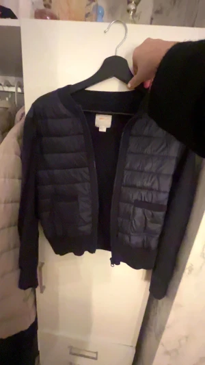 Svart quiltad pufferjacka med dragkedja - Snygg svart pufferjacka med quiltade paneler framtill och mjuka stickade ärmar. Jackan har rund hals, praktisk dragkedja och en liten ficka med ribbad kant. Perfekt för dig som gillar en clean och modern look.