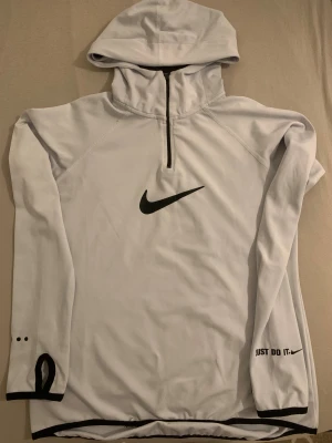 Vit Nike hoodie med half zip och huva - Nike hoodie i vit med svart logga och texten 'Just Do It.' på ärmen. Tröjan har huva, half zip och svarta detaljer vid muddar och nederkant. Tillverkad i mjuk polyester med meshfoder i huvan, perfekt för träning eller chill. Tumhål på ärmarna för extra komfort.