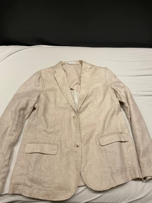 Beige linnekavaj med klassisk passform 164cm - Snygg beige kavaj från H&M i linne med två knappar framtill och klassiska fickor med lock. Perfekt för dig som gillar en stilren look med naturliga material. Passar dig som vill ha en lätt och luftig kavaj till varmare dagar.