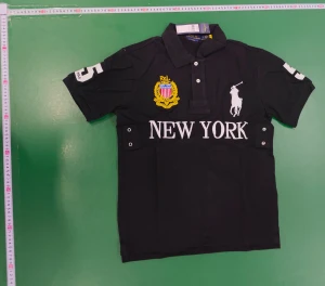 Svart Polo Ralph Lauren New York piké - Svart pikétröja från Polo Ralph Lauren med broderad gul och vit logga, stor vit text 'NEW YORK' på bröstet och siffran 5 på ärm och rygg. Klassisk krage, kort ärm och knappar framtill. Perfekt för dig som gillar sportig och stilren look.