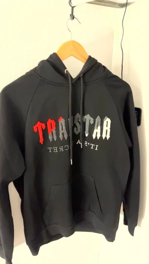 Svart Trapstar hoodie med tryck - Svart hoodie från Trapstar i storlek M med huva och snörning. Framsidan har ett stort broderat Trapstar-tryck i rött, grått och vitt. Klassisk känguruficka framtill och mjukt material som känns skönt mot huden. Perfekt för dig som gillar streetwear.