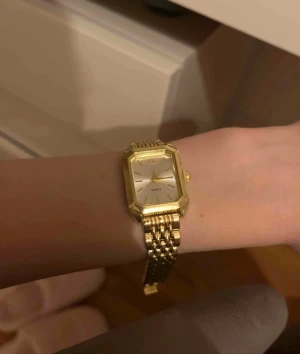 Guldig fyrkantig klocka med länkarmband - Säljer en stilren klocka med fyrkantig urtavla i silver och guldig ram. Klockan har ett klassiskt guldigt länkarmband och minimalistisk design utan siffror, perfekt för dig som gillar en clean look.
