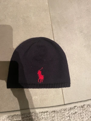 Marin blå mössa från Polo Ralph Lauren - Marin blå mössa från Polo Ralph Lauren i 100% ull. Klassisk rund form med röd broderad logga framtill som ger en snygg kontrast. Perfekt för dig som vill ha en stilren och enkel mössa med exklusiv känsla. Passar dig som gillar ikoniska märken och clean design.