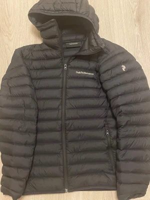 Svart dunjacka Peak Performance - Svart dunjacka från Peak Performance med huva och broderad logga på bröstet. Jackan har quiltad design, dragkedja framtill och två sidofickor med dragkedja. Perfekt för kalla dagar och har en stilren look som funkar till allt.
