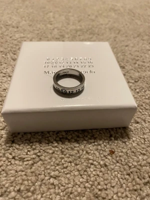 Maison Margiela silverring med siffror - Cool och chunky silverring från Maison Margiela, tillverkad i Italien av äkta 925 sterling silver. Ringen har ett unikt designmönster med svarta siffror runt hela bandet och den klassiska Margiela-logon. Perfekt statement piece för dig som gillar modern och edgy stil.  19mm inre diameter