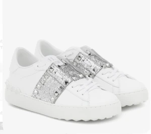 Valentino Rockstud glitter sneakers - Säljer ett par vita Valentino Rockstud sneakers med silverglittriga band och nitar över ovansidan. Skorna har snörning, platt sula och är tillverkade i skinn. De är ganska använda vilket syns på bilderna därav nedsatt pris, nypris 8500kr.