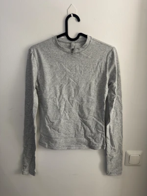 Grå tight långärmad tröja från SKIMS - En enkel och stilren grå långärmad tröja från skims. Tröjan har rund hals och är gjord i mjuk bomull med lite elastan för extra stretch. Perfekt att matcha med jeans eller under en jacka. Snygg basic som funkar året runt.