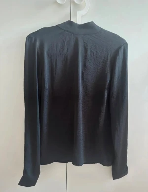 Svart blus med öppning i ryggen - Snygg svart blus med långa ärmar och halvpolo. V-ringad öppning bak med knytband. Blusen är tillverkad i glansigt satinliknande material. Perfekt för dig som gillar en stilren look med en twist