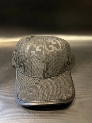 Svart keps från med GG-mönster - Snygg svart keps med det klassiska GG-mönstret ton-i-ton över hela kepsen. Tillverkad i bomull med detaljer i skinn. Justerbar rem baktill i svart läder med guldfärgad spänne. Perfekt för dig som gillar exklusiva accessoarer.