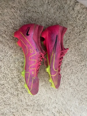 Nike rosa fotbollsskor med snörning - Säljer ett par Nike fotbollsskor PRO i en riktigt fet rosa färg med gula detaljer och sula. Skorna har snörning och är gjorda i syntetmaterial med stickad ovandel. Swoosh-loggan syns tydligt på sidan och ovansidan. Perfekt för dig som vill sticka ut på planen.