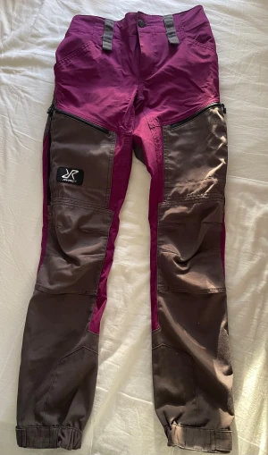 RevolutionRace byxor  - Säljer ett par täckbyxor från RevolutionRace i snygg rosa och grå färg. Byxorna har elastisk midja. Perfekta för utomhusaktiviteter. 