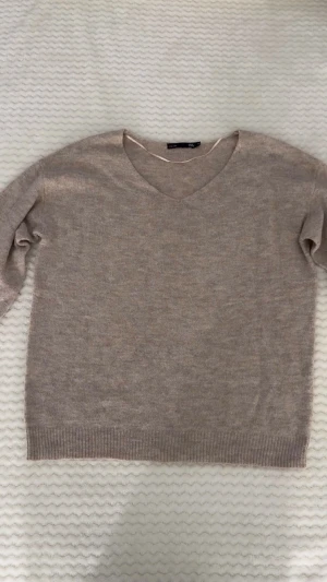 Beige v-ringad stickad tröja 157 - Mysig beige stickad tröja från 157 med v-ringad hals och långa ärmar. Tröjan har ribbade muddar vid ärmslut och nederkant, perfekt för lager-på-lager under hösten och vintern. Enkel och stilren design som funkar till jeans eller kjol.