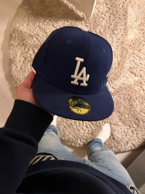 Blå LA Dodgers keps från New Era - Säljer en blå New Era 59FIFTY keps med LA Dodgers-logga i vitt framtill. Klassisk flat peak och broderad detalj på toppen. Kepsen är gjord i polyester och har en snygg, sportig look som passar perfekt till streetwear. Ikonisk modell för dig som gillar baseballstil.