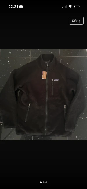 Svart fleecejacka från Patagonia - Svart fleecejacka från Patagonia med hög krage och hel dragkedja framtill. Jackan har två sidofickor med dragkedja och ett litet broderat logomärke på bröstet. Mjuk teddystruktur och normal passform, perfekt för kyliga dagar.