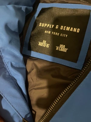 Blå pufferjacka Supply & Demand - Snygg blå pufferjacka från Supply & Demand med huva och coolt märke på ärmen. Jackan har dragkedja framtill och quiltad design som ger en schysst streetstil. Perfekt för dig som gillar att sticka ut med en fet jacka i vinter.