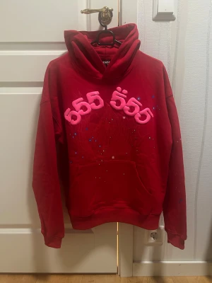 SP5R Hoodie - Cool röd hoodie från SP5R i storlek S med stor huva och magficka. Framsidan har ett fett 3D-tryck med rosa siffror '555' och stjärnor, samt ett spindelnätsmönster i bakgrunden. Perfekt för dig som gillar unika streetwear-plagg.