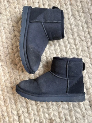 Marinblå UGG boots i mocka, storlek 36 - Mysiga marinblå UGG boots i klassisk modell ”Mini II” med rund tå och platt sula. Utsidan är i mjuk mocka och insidan är fodrad med fluffigt material som håller fötterna varma. Diskret UGG-logga baktill och grov yttersula för extra grepp. Perfekt för kalla dagar. Nypris omkring 1700kr. Köpta på zalando. 