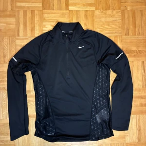 Svart Nike Dri-Fit träningströja S - Svart långärmad träningströja från Nike med Dri-Fit-teknologi. Tröjan har half zip framtill, diskreta mönstrade paneler på sidorna och reflekterande detaljer på ärmarna. Perfekt för löpning eller gym. Snygg sportig look med liten Nike-logga på bröstet.