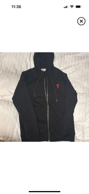 Svart hoodie från Ami Paris med rött hjärta - Svart hoodie från Ami Paris med dragkedja, huva och snörning. På bröstet finns ett broderat rött hjärta med bokstaven A. Tröjan har långa ärmar och ribbade muddar. Perfekt för dig som gillar stilrena streetwear-plagg med en twist.