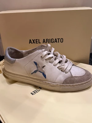 Axel Arigato vita sneakers med broderad fågel - Tjena säljer  ett par vita sneakers från Axel Arigato med broderad fågel i blått och grått på sidan.  Skorna har en liten reva i mockan på ena skon annars är skon i hyffsar bra skick. Kvitto finns på sista bilden. 