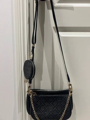 Svart axelväska från Silvio Tossi med kedja -  svart axelväska från Silvio Tossi  med quiltad yta och präglat logomönster. Väskan har guldfärgade kedjedetaljer, dragkedja och en rund liten coin pouch som hänger på axelremmen. Kan användas på flera olika sätt 