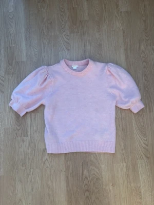 Ljusrosa stickad tröja med puffärm - Supersöt ljusrosa stickad tröja med korta puffärmar och ribbade muddar. Tröjan har rund halsringning och är i ett mjukt material som känns skönt mot huden. Perfekt för dig som gillar en feminin och trendig stil. 