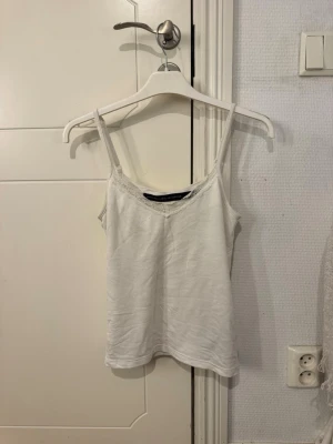 Vit linnetopp med spetsdetalj - En enkel och stilren vit linnetopp med smala axelband och diskret spetskant vid ringningen. Perfekt att matcha med jeans eller kjol för en fräsch look. Mjuk och bekväm känsla tack vare bomullsmaterialet.