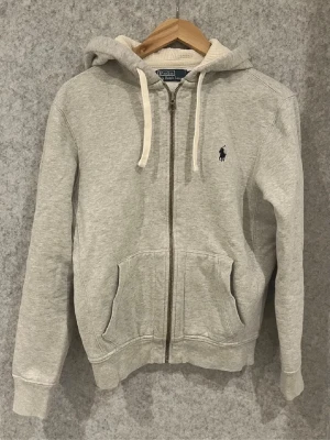 Grå hoodie från Polo Ralph Lauren - Grå zip-hoodie från Polo Ralph Lauren med vit dragsko i huvan och klassisk broderad logga på bröstet. Mjuk bomullskänsla, kängurufickor och ribbade muddar. Perfekt för en chill och stilren look.