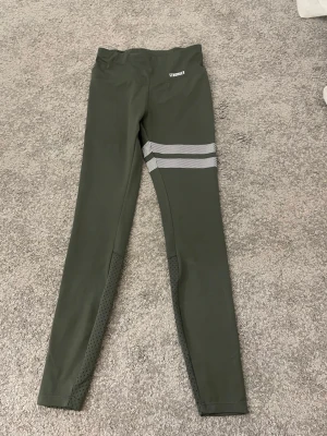 Gröna leggings från Stronger - Mörkgröna leggings från Stronger med två vita diagonala ränder på ena benet och ventilerande meshdetaljer vid vaderna. Snygg och sportig design som passar perfekt till träning. Materialet känns stretchigt och bekvämt.