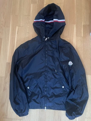 Marinblå vindjacka från Moncler - Snygg marinblå vindjacka från Moncler med huva och vit-röd rand på huvan. Jackan har dragkedja framtill, två fickor med knappar och Moncler-logga på ärmen. Tillverkad i lätt polyester, perfekt för blåsiga dagar.