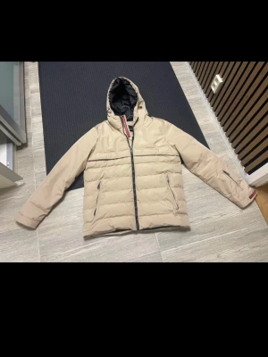 Beige dunjacka med huva - Snygg beige dunjacka med huva och svarta detaljer. Jackan har dragkedja framtill, två sidofickor med dragkedja och en quiltad design. Perfekt för kalla dagar och har en modern, clean look. Köpt här, osäker på om den är äkta, därav priset