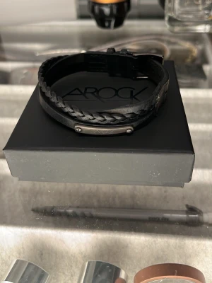 Svart läderarmband från Arock - Stilrent svart armband från Arock i äkta läder med både slät och flätad rem. Armbandet har en metallplatta med Arock-logga och justerbart spänne. Perfekt accessoar för en modern look. Priset är inte satt än så kom med bud men utgå från 300kr!☺️