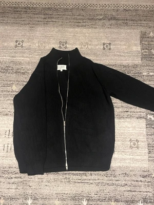 Svart ziptröja från Maison Margiela - Säljer en helt ny svart ribbstickad ziptröja från Maison Margiela. Tröjan har hög krage, hel dragkedja framtill och är tillverkad i ett mjukt, stickat material. Perfekt för lager-på-lager och en clean look. OBS… kan tänka mig gå ner nån hundra lapp vid snabbaffär!