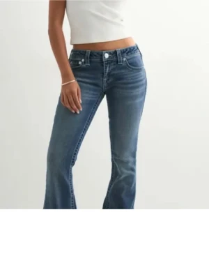 True religion  - Säljer ett par blå bootcut jeans med snygga broderade bakfickor och klassisk femficksdesign. Jeansen har låg midja och är tillverkade i mjukt denimtyg som sitter skönt. Perfekta för dig som gillar en avslappnad men trendig look.