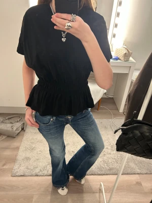 Svart topp med volang och resårmidja - Snygg svart topp med korta ärmar och markerad resår i midjan som ger en fin siluett. Toppen har volang nedtill och är i ett mjukt, tunt material som känns skönt mot huden. Perfekt att styla med jeans eller kjol för en trendig look. Säljer denna topp då den bara har hängt i min garderob och har kommit till användnings ca 1-3 gånger. Djur och rökfritt hem, Svart topp i storlek XS från Lager 157! Nypris: 150kr. ☺️(Säljs ej längre i butik.)