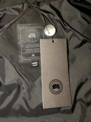 Svart dunjacka från Canada Goose - Säljer en svart dunjacka från Canada Goose i modellen Fusion. Jackan har huva, ribbade muddar och klassisk patch på ärmen. Perfekt för kalla dagar och riktigt snygg till vinterlooken.