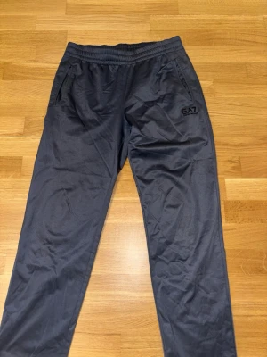 Svarta mjukisbyxor från EA7 Emporio Armani - Snygga svarta mjukisbyxor från EA7 Emporio Armani med elastisk midja och två sidofickor. Byxorna har en diskret logga på vänster lår och är tillverkade i ett mjukt och lätt material som känns skönt mot huden. Perfekta för chill eller träning.