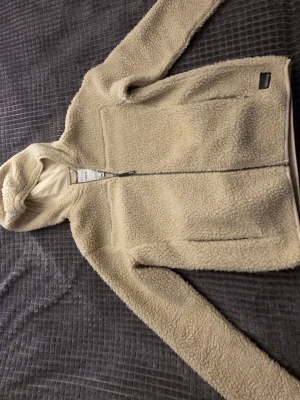 Beige teddyjacka med huva  - Mysig beige teddyjacka med huva och dragkedja framtill. Jackan har två sidofickor och är tillverkad i fluffigt teddy-material som håller dig varm under kyliga dagar. Perfekt för dig som gillar en avslappnad och trendig stil.