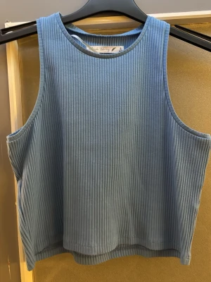 Blå ribbad ärmlös linnetopp - Snygg blå ribbad linnetopp med rund halsringning och avslappnad passform. Toppen är ärmlös och har en croppad längd som passar perfekt till höga jeans eller kjol. Enkel men trendig design som är lätt att matcha.