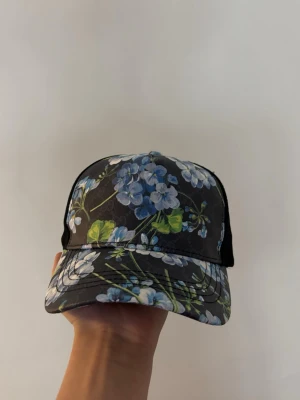 keps med blått blommönster - Snygg keps  med svart bas och blått blommönster. Kepsen har justerbar rem baktill och klassisk böjd skärm. Snygg logga och detaljer i svart och vitt. Perfekt för att lyfta din outfit.