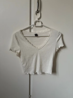 Vit ribbad croppad topp med spets - Söt vit croppad topp med ribbad struktur och v-ringning. Korta ärmar och spetsdetalj längs halsringningen ger en extra touch. Perfekt att styla med jeans eller kjol för en fräsch look.