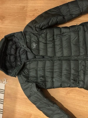 Grön dunjacka från Arc'teryx - Snygg svart dunjacka från Arc'teryx med huva och dragkedja framtill. Jackan har quiltad design och två sidofickor med dragkedja. Perfekt för kyliga dagar och enkel att matcha med olika outfits. Äldre modell Cerium SV
