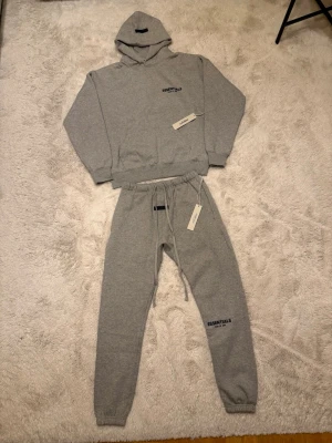 Grå Essentials Fear of God hoodie och mjukisbyxor - Snygg grå hoodie och mjukisbyxor från Essentials Fear of God med svart logga på bröstet och ryggen och under knät. Klassisk passform med huva och ribbade muddar. Perfekt för en avslappnad streetwear-look. Helt oanvänd och Storlek S.
