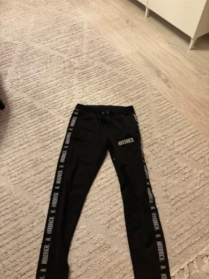 Svarta mjukisbyxor från Hoodrich - Svarta mjukisbyxor från Hoodrich med logga tryckt på vänster ben och längs båda sidorna. Byxorna har snörning i midjan och smal passform. Perfekta för chill eller streetwear-stil.