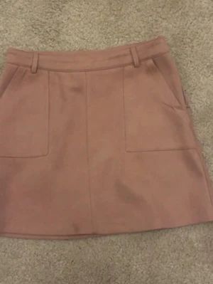 Rosa A-linjeformad kjol med fickor - Söt rosa kjol i A-linjeform med två stora framfickor och hällor för skärp. Kjolen har en enkel och stilren design, perfekt för att matcha med olika toppar. Materialet ser ut att vara mjukt och något tjockare, vilket ger en snygg struktur.