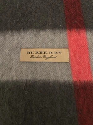 Rutig halsduk från Burberry - Snygg halsduk från Burberry i klassiskt rutigt mönster med grå, svart, vit och röd färg. Tillverkad i mjukt material och har fransar i ändarna. Perfekt accessoar för att lyfta din outfit med en tidlös och lyxig känsla.