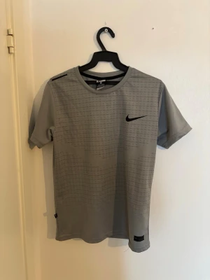 Grå Nike t-shirt med tryck - Snygg grå t-shirt från Nike med diskret mönster och svart logga på bröstet. T-shirten har rund halsringning och korta ärmar. Tillverkad i ett lätt och funktionellt polyestermaterial som passar perfekt till träning.