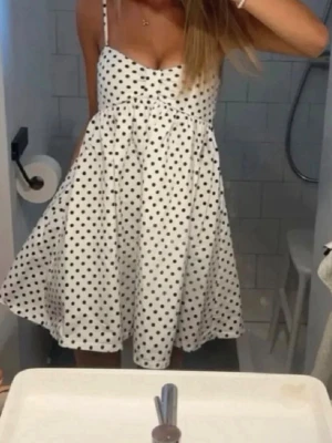 Vit prickig kortklänning med smala band - Söt vit kortklänning med svarta prickar och smala axelband. Klänningen har en lätt utställd kjol och markerad byst, vilket ger en feminin siluett. Perfekt för varma dagar och enkel att styla med accessoarer.