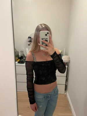 Svart mesh offshoulder topp - Trendig svart offshoulder topp i mesh med långa ärmar och rynkade detaljer framtill och på ärmarna. Croppad modell som ger en snygg och edgy look. Perfekt att styla med jeans eller kjol för en cool outfit.